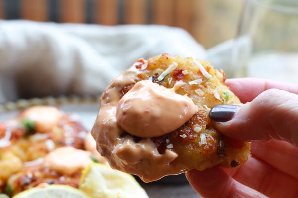 A person's hand holding a golden-brown coconut shrimp cakes topped with a dollop of spicy aioli sauce.