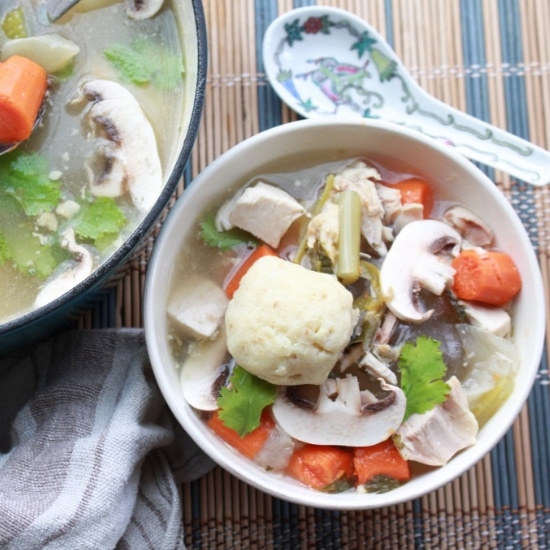A white bowl of matzo ball soup with veggies on top and a soup spoon on the side with a pot of soup in the background placed on top of a bamboo placemat.