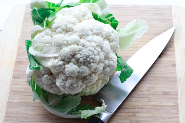 A head of white cauliflower on top of a wooden cutting board with chef's knife on the side.