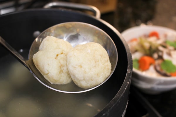 A soup ladle with two fluffy matzo balls over a pot of water and a bowl of matzo ball soup in the background.