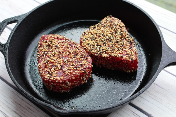 Two tuna steaks dredged in sesame seeds in a cast iron skillet ready to be seared placed on top of a white wooden board.