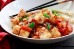 Crispy tofu cubes in a sweet and sour sauce in a white bowl with black chopsticks on the side.