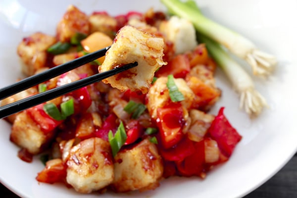 crispy tofu cubes in a white bowl tossed in a sweet and sour sauce with black chopsticks