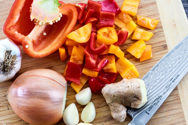 Colorful red and yellow bell peppers, garlic cloves, onion, and ginger on a wooden cutting board.