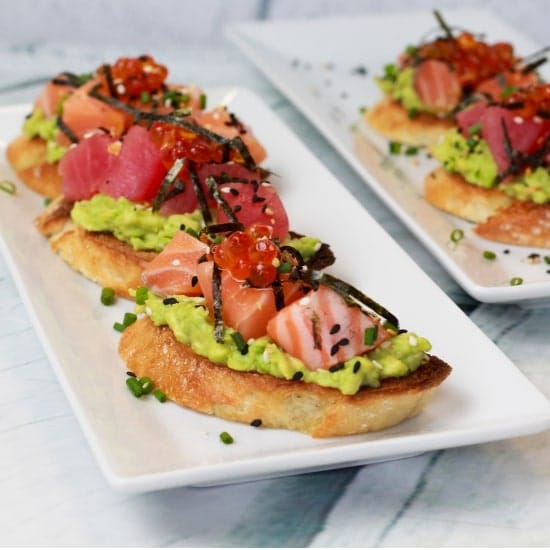 Sushi-grade salmon and tuna are nestled on top of avocado toast presented on a two white plates with a blue napkin on the side.