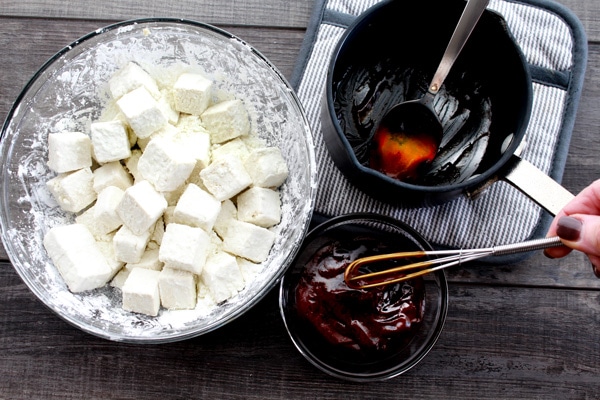 Cubed tofu dredged in corn starch in a glass bowl with a sauce in a bowl and in a small pot on the side on top of a wooden board.