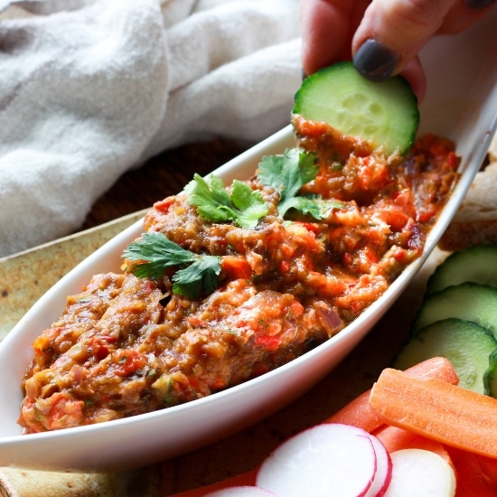 Roasted Chinese eggplant dip in a white boat-shaped bowl with fresh veggies and sliced bread on the side and a hand dipping a cucumber into the dip.