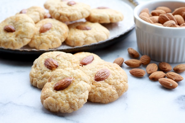 A stack of Chinese almond cookies on a round plate with a bowl of almonds on the side and four cookies in the front.