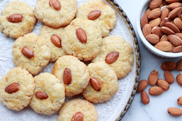Chinese almond cookies piled on a round white plate with bowl of almonds on the side and extra cookies in front on top of a marble surface.
