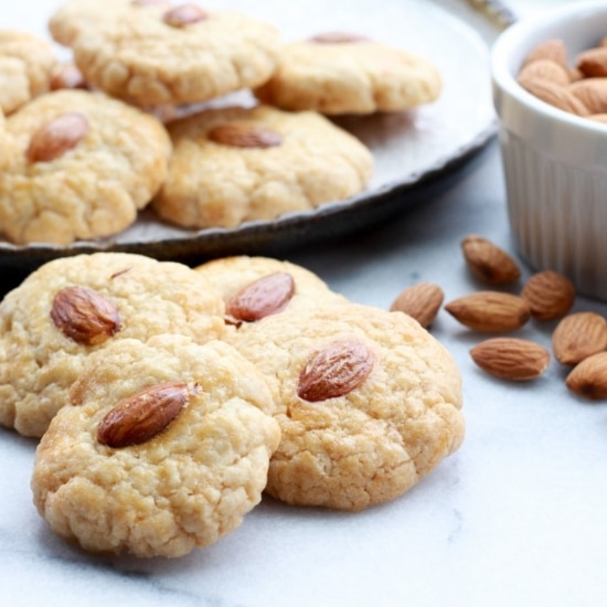 Chinese almond cookies piled on a round white plate with bowl of almonds on the side and extra cookies in front on top of a marble surface.