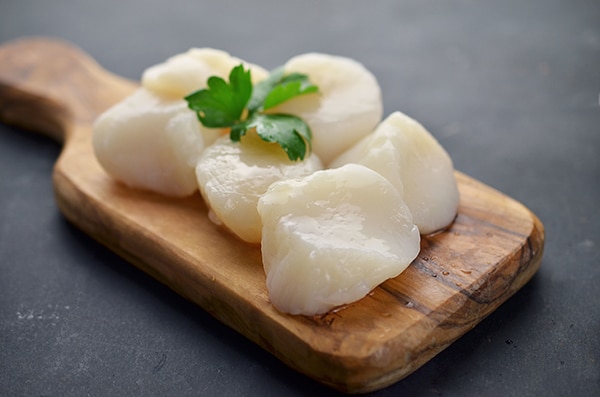 Raw sea scallops topped with parsley on a small wooden cutting board.