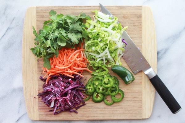 fresh chopped veggies being prepared for Asian slaw with Nuoc Cham dressing on top of a wooden board with a chef's knife on the side.
