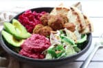 falafel balls in a bowl with beet hummus, slaw, red onions, and avocado on a white board and a fork on the side