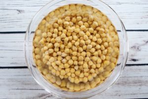 chickpeas soaking in water in a glass bowl on top of a white wooden board