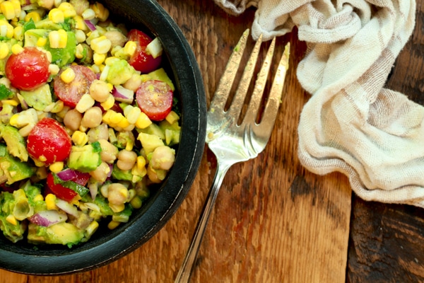 a vibrant corn and avocado salad in a black wooden bowl with a large silver fork and linen napkin the side on top of a wooden board