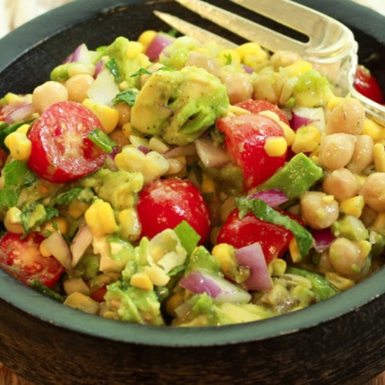 a vibrant corn and avocado salad in a black wooden bowl with a large silver fork and linen napkin the side on top of a wooden board