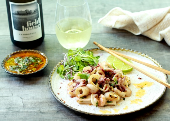 pan seared calamari on a white plate with chopsticks, a side of nuoc cham sauce, and glass of white wine on a wooden board