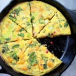 a roasted butternut squash and baby bok choy frittata with one slice missing in a cast iron pan on top of a gray board