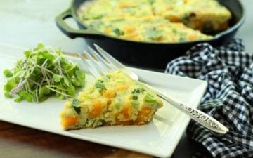 a slice of roasted butternut squash and baby bok choy frittata on a white plate with fresh herbs and a silver fork and checkered napkin on the side and a frittata in a cast iron pan in the background