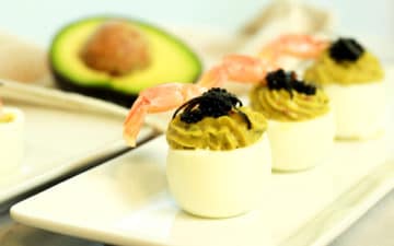 avocado deviled eggs topped with fresh shrimp and caviar stacked on a white plate with a halved avocado in the background.