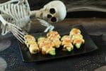 Scary Halloween Jalapeño Popper Mummies on a black plate with a skeleton sitting behind.