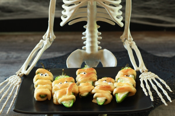 Halloween Jalapeño Popper Mummies on a black plate with a skeleton sitting behind.