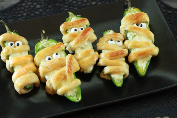 jalapeńo popper mummies encased in phylo dough decorated with candy eyes on top of a black plate.