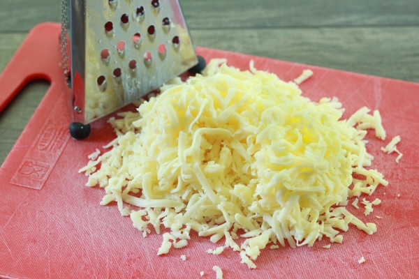 Shredded white cheese on a red cutting board with a cheese grater alongside
