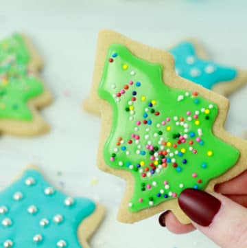 A woman's hand holding up a decorated green Christmas tree sugar cookie with other cookies in the background.