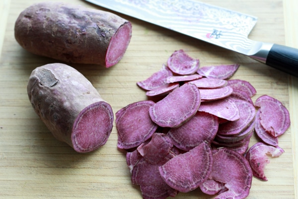 purple sweet potatoes slices and halved sweet potatoes on a wooden cutting board with a chef's knife on the side