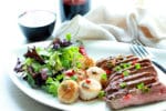 Seared scallops and grilled flank steak on a white plate with a bottle and glass of red wine