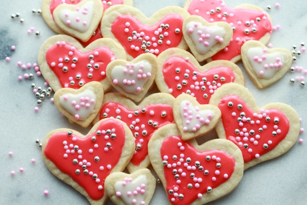 Heart-shaped sugar cookies in various sizes with pink and white glaze and decorative sprinkles stacked on top of a marble surface.