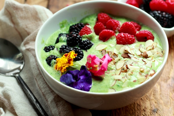 a vibrant green smoothie bowl topped with raspberries, blackberries, almonds and edible flowers on top of a wooden board with fresh berries and an avocado on the side