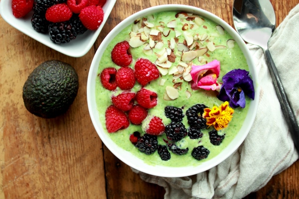 a vibrant green smoothie bowl topped with raspberries, blackberries, almonds and edible flowers on top of a wooden board with fresh berries, an avocado, and a spoon on the side.