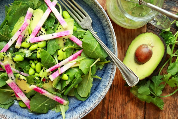 avocado-edamame kale salad in a blue bowl with a fork and avocado and dressing on the side
