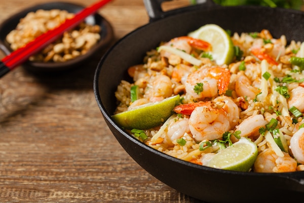 shrimp fried rice in a large black wok on a wooden board with a small bowl of peanuts and red chopsticks behind