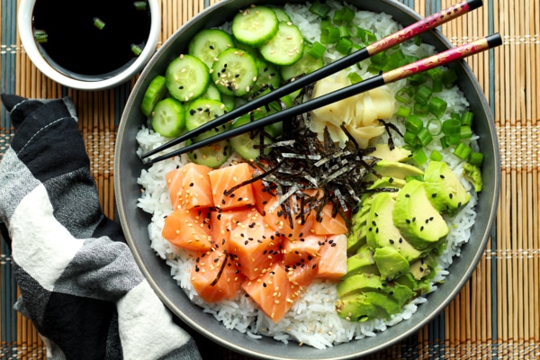Salmon sushi bowl featuring chunks of sushi-grade salmon, sushi rice, sliced avocados, cucumbers, and seaweed strips on a bamboo mat with chopsticks.