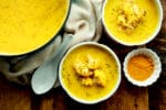 vibrant cauliflower soup in two white bowls on a wooden board with turmeric powder along side