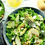 A bowl of arugula salad topped with Asian pears and avocados with dressing on the side