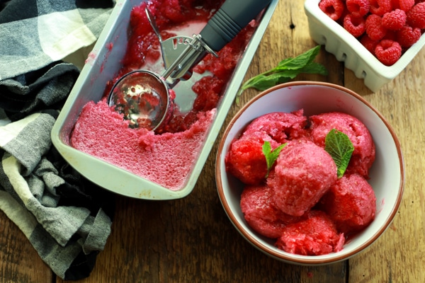 a bowl of raspberry sorbet in a white bowl on a wooden board with a metal pan filled with sorbet and fresh raspberries on the side