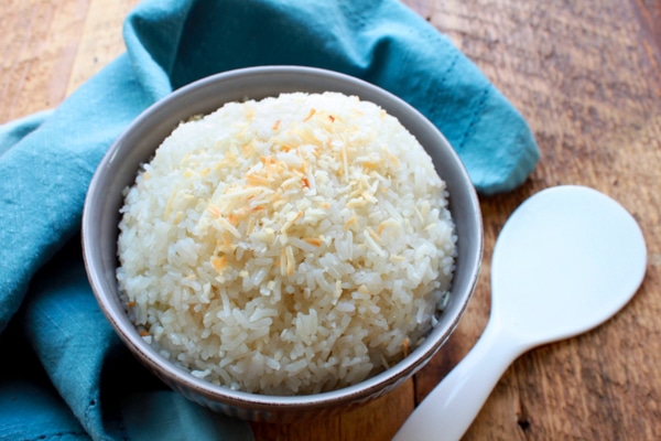 A gray bowl filled with steamed white rice topped with coconut flakes, a white spoon and blue napkin on the side, on top of a wooden board