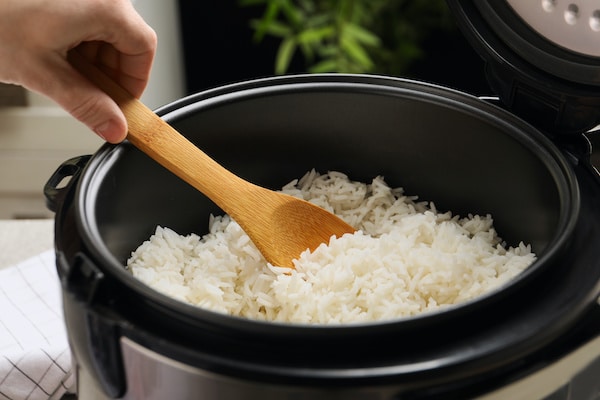 A woman holding a wooden spoon scooping white rice from a rice cooker