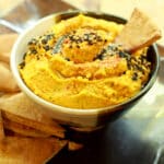 a small bowl of roasted carrot and ginger hummus with a pita chip inserted and pita chips on the side