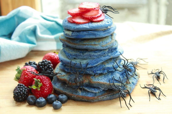 stacks of blue pancakes with spiders crawling up and berries on the side on top of a wooden board