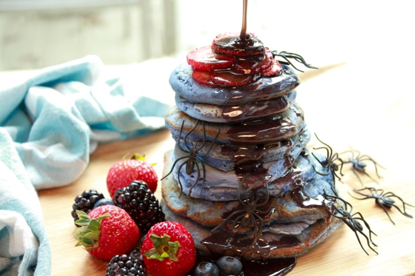 a stack of blue pancakes with black syrup dripping down the stack with vibrant berries on the side and plastic spiders crawling up the stack
