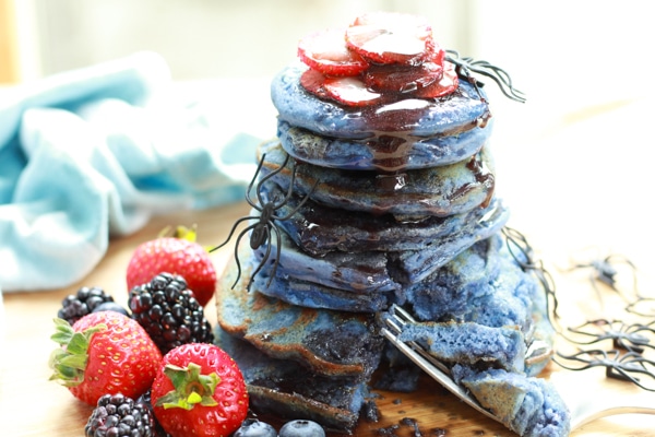 a stack of blue pancakes with black syrup dripping down the stack with vibrant berries on the side and plastic spiders crawling up the stack