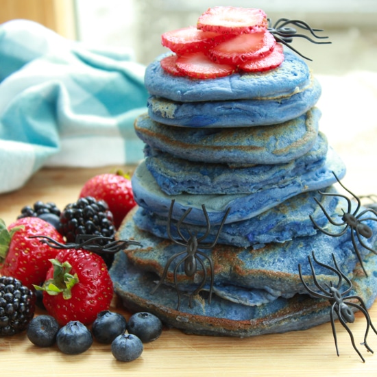 a stack of blue pancakes on a wooden board with vibrant berries on the side and plastic spiders crawling up the stack