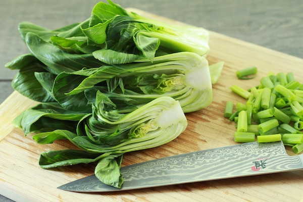 Baby bok choy on a wooden cutting board with a chef's knife and chopped scallions on the side