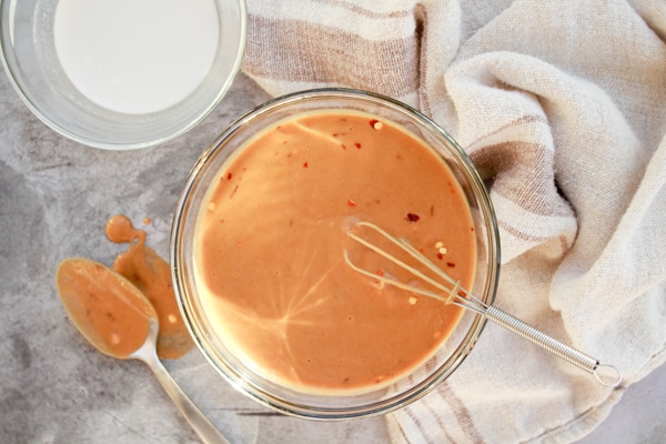 Peanut sauce inside a clear glass bowl with a whisk inserted inside the bowl and a spoon and napkin on the side