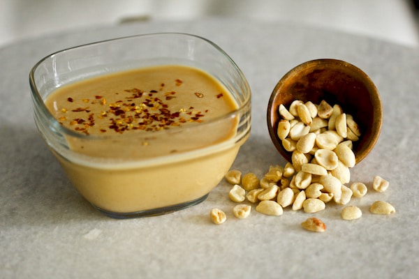 Peanut sauce inside a clear glass bowl with loose raw peanuts on a gray board.